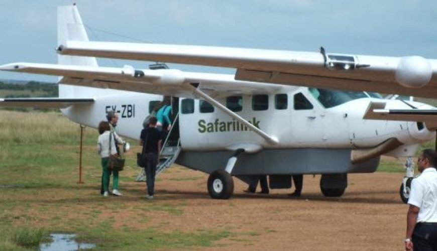 Small aircraft landing on remote airstrip with safari vehicles and wildlife in the background